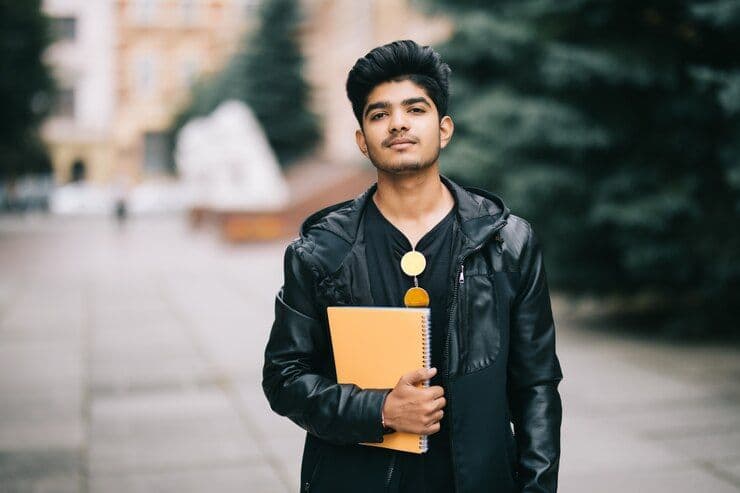Indian student standing in front of a university in Germany holding books and a backpack.jpg